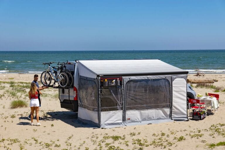 Camper op het strand met uitgerolde luifel en wanden eromheen, twee mensen ervoor