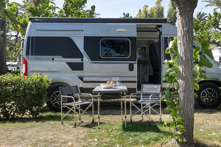 Twee campingstoelen en een tafel naast een buscamper met open schuifdeur op een camping