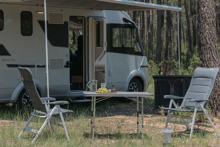 Twee grijze Crespo campingstoelen met hoofdsteun bij een tafel naast een integraalcamper aan de rand van een bos