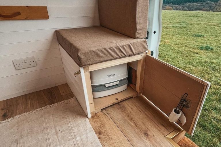 Porta Potti in een buscamper: het chemische toilet staat achter een houten deurtje onder de zitbank, met de schuifdeur open op een weiland