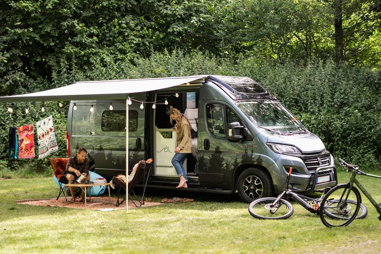 Scheidingstoilet camper: buscamper met uitgedraaide luifel op een camping, met twee mensen aan een tafeltje en fietsen ervoor